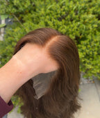 Person holding a brown wig against a blurred green outdoor background