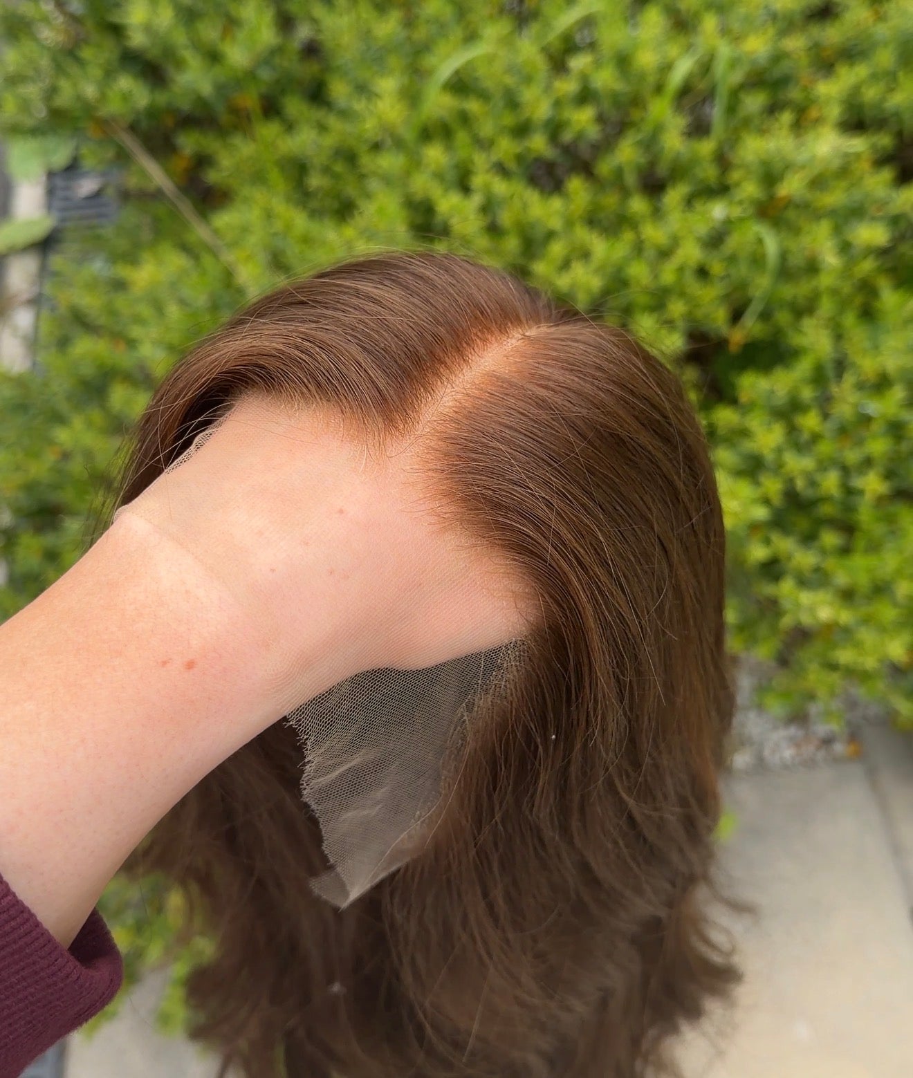 Person holding a brown wig against a blurred green outdoor background