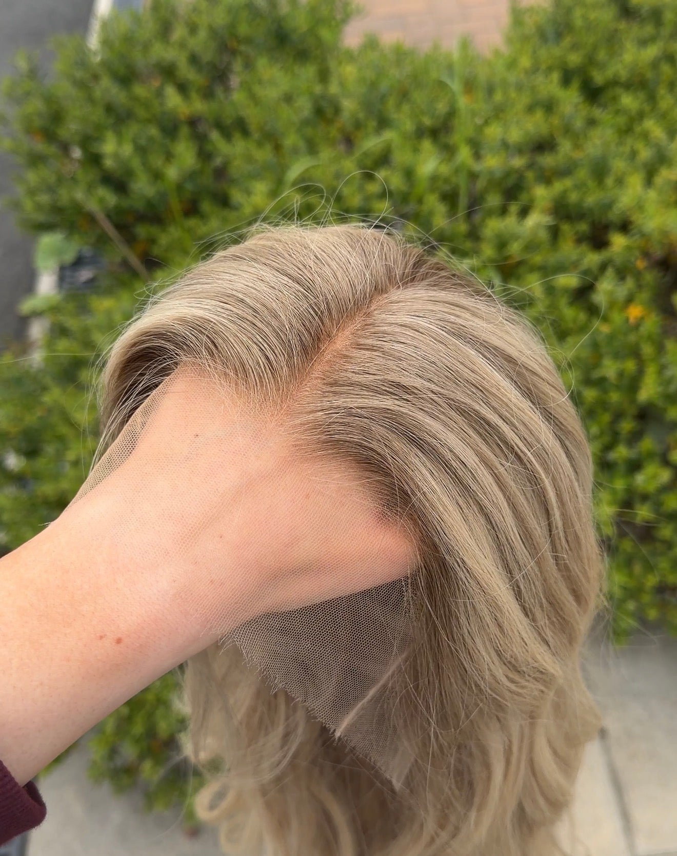 Person with a blonde wig held up against a blurred green outdoor background